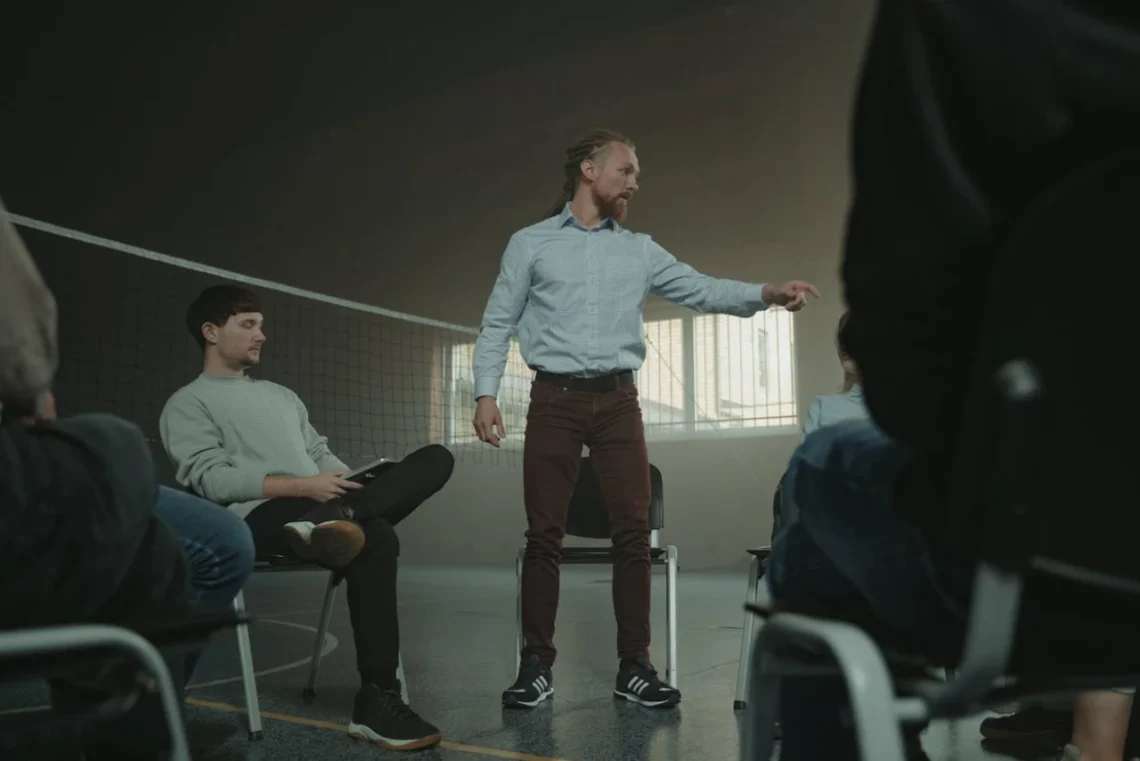 Mand står i sportshal med kreds af mennesker som i coaching.